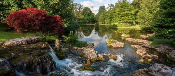 The view from the waterfall in St. Fiachra's Garden showing a bright red Japanese maple in the foreground, the lake and rocks in the middle ground, and green trees and blue sky in the background.