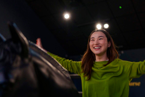 A young woman sitting on the racing simulator in the Irish Racehorse Experience celebrates her win with a smile and her arms wide.